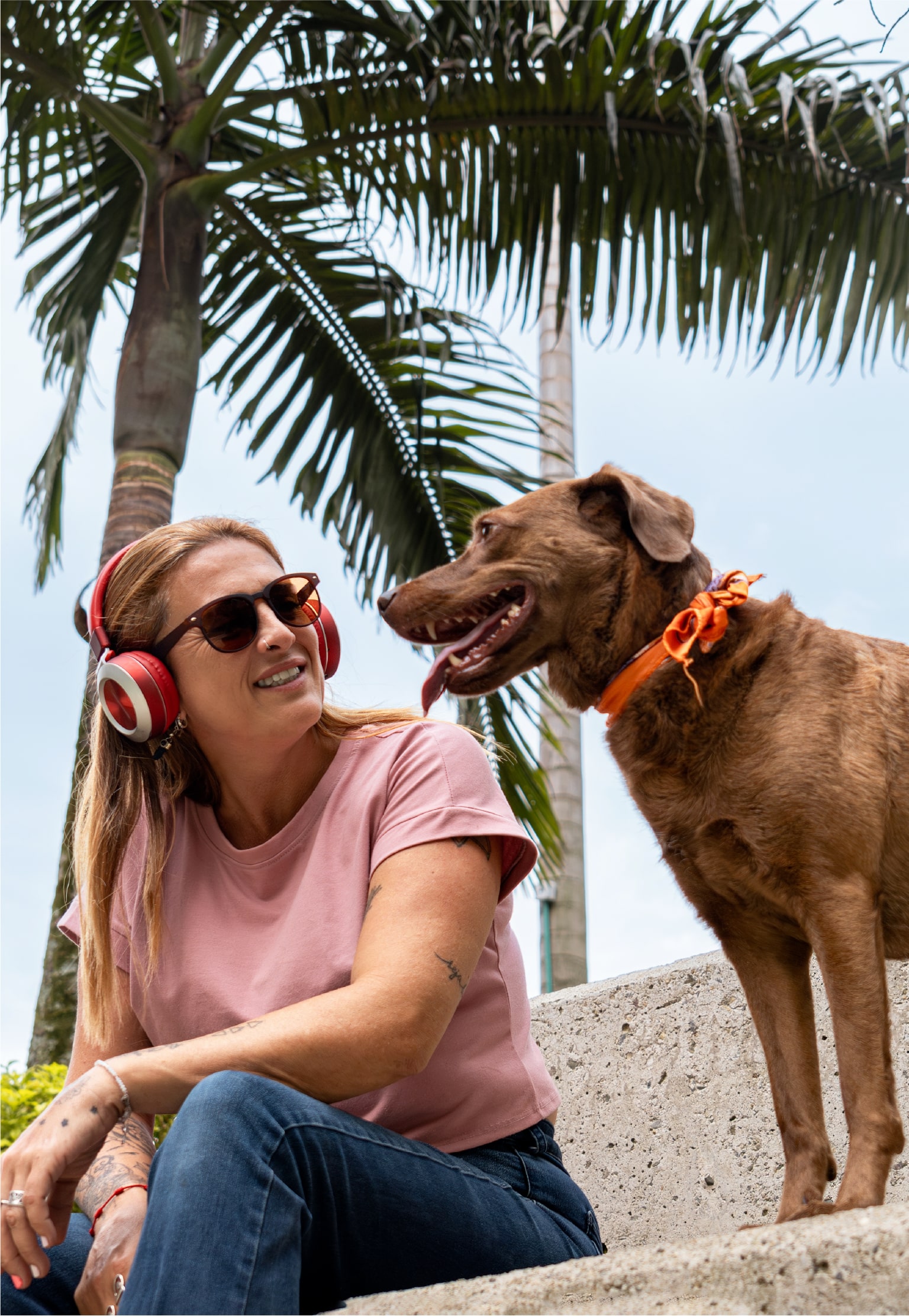 Woman wearing headphones smiles at a dog with an orange collar against a palm backdrop.